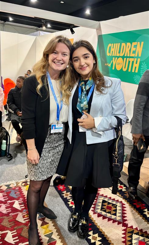 Two women standing together and smiling at an event, with a banner in the background reading "Children and Youth.