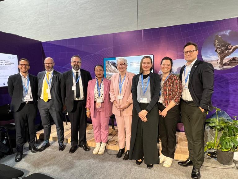 A group of eight people in formal attire standing in a line indoors, with a purple backdrop and a screen behind them.