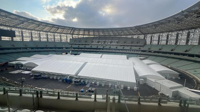 Large open-air stadium with a white tented structure covering most of the field area. Overcast sky visible through the stadium opening.