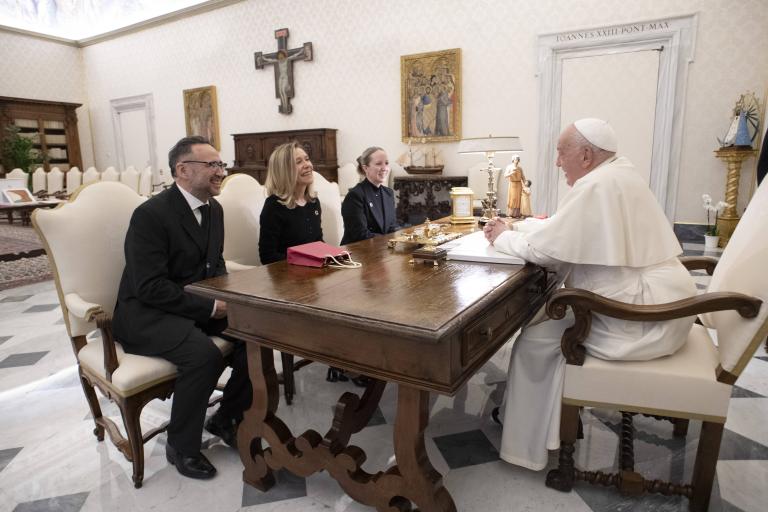 A group of three people sitting across from the Pope at a wooden table in a formal room with religious decor.