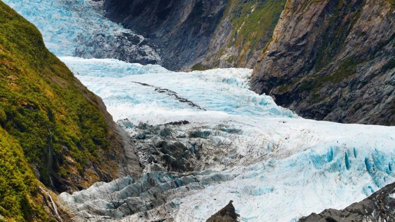 A glacial valley with icy blue glacier flowing between rocky, green-covered mountains.