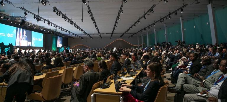 A large conference room filled with seated attendees facing a stage with screens displaying presentations.