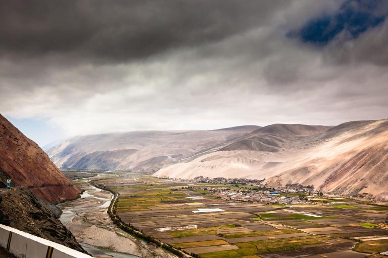 A scenic view of a river winding through agricultural fields and a small settlement, surrounded by rolling hills under a cloudy sky.