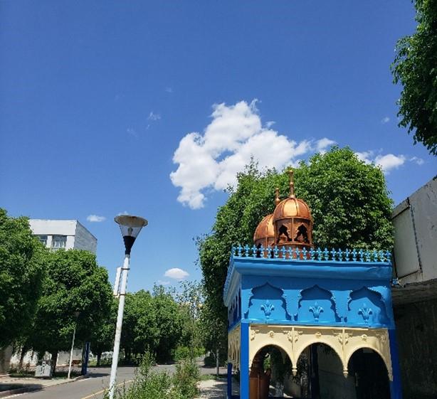 A blue and yellow structure with a dome, set against a clear sky with white clouds, surrounded by green trees and a streetlight.