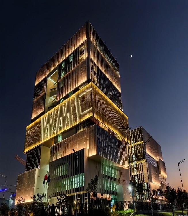 A modern, multi-story building with illuminated façades at night. The structure features grid-like patterns and warm lighting, set against a dark sky with a visible crescent moon.