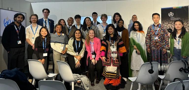 A diverse group of people stand together, some wearing cultural attire, in a conference room with chairs and a banner in the background.