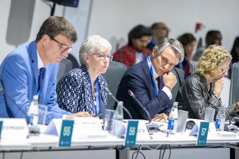 A group of people in a formal setting, seated at a table with microphones, nameplates, and water bottles, engaged in discussion.