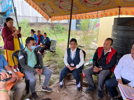 A group of people, some seated under a canopy, are engaged in conversation. A woman in a uniform is at the centre, and others are attentively listening.