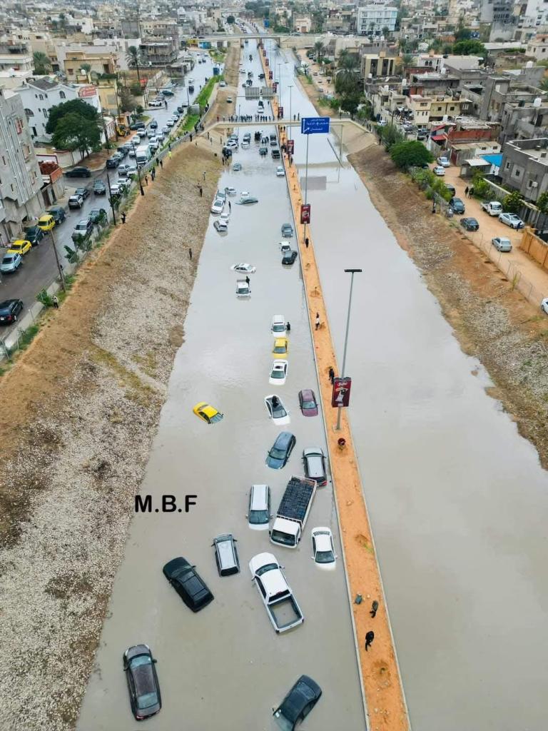 A flooded street with partially submerged cars lined along both sides, surrounded by buildings in an urban area.