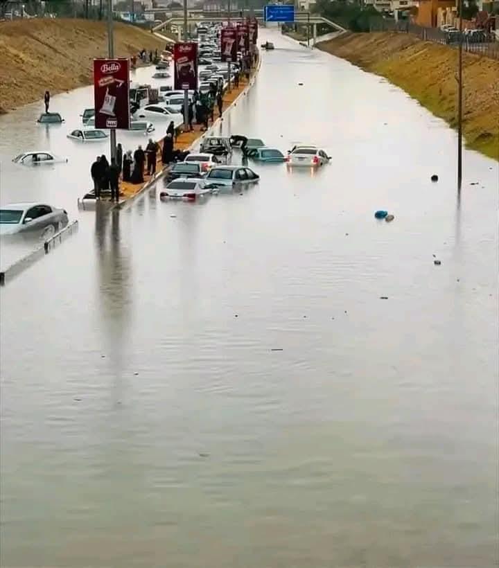 Flooded street with submerged cars and people standing on a raised walkway, surrounded by water, and traffic stopped.