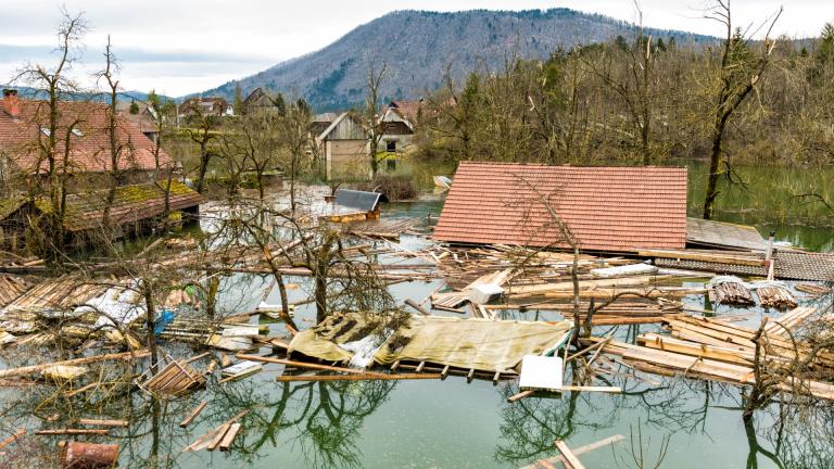 Flooded residential area with debris and damaged houses, partially submerged under water. Trees and materials are scattered. Mountains are visible in the background.