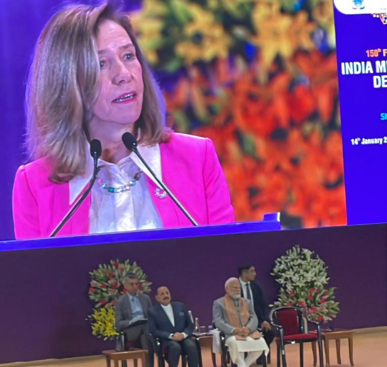 A woman speaks at a podium with a large screen displaying her image. Three men, including one wearing a shawl, sit on stage with floral arrangements in the background.