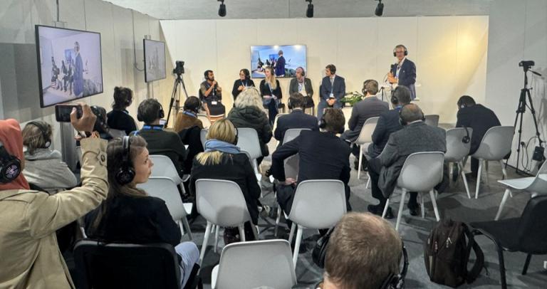 Audience seated in a conference room listens to a panel discussion. Panelists are seated at the front, and several attendees wear headphones. Cameras are set up for recording.