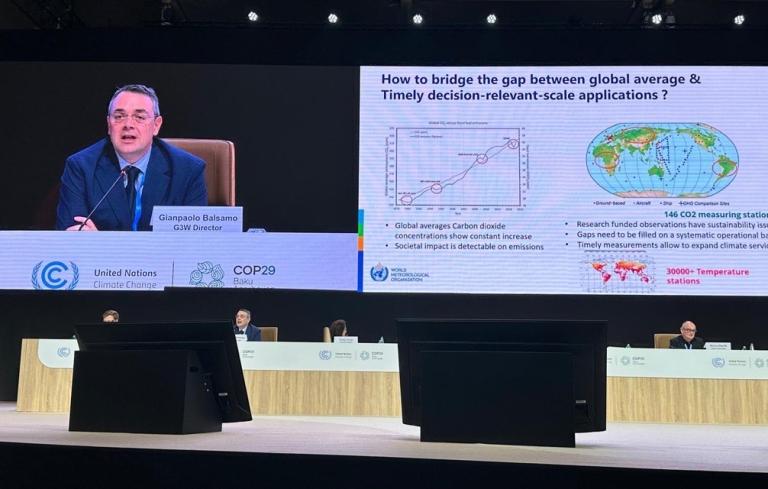 A speaker presents data on climate change at a conference with graphs on a large screen. Participants are seated at a table in front.