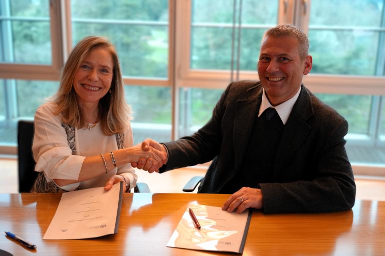 Two people are sitting at a table, shaking hands over signed documents. There are pens placed on the papers. The background shows large windows with a view of greenery.