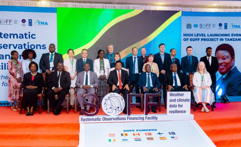 A group of people dressed in formal attire poses on a stage at an event. A "SOFF" sign and banners about weather and climate data are displayed.