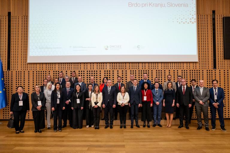 A group of people in formal attire stands on a stage with a wooden backdrop and a Slovenian location shown above.