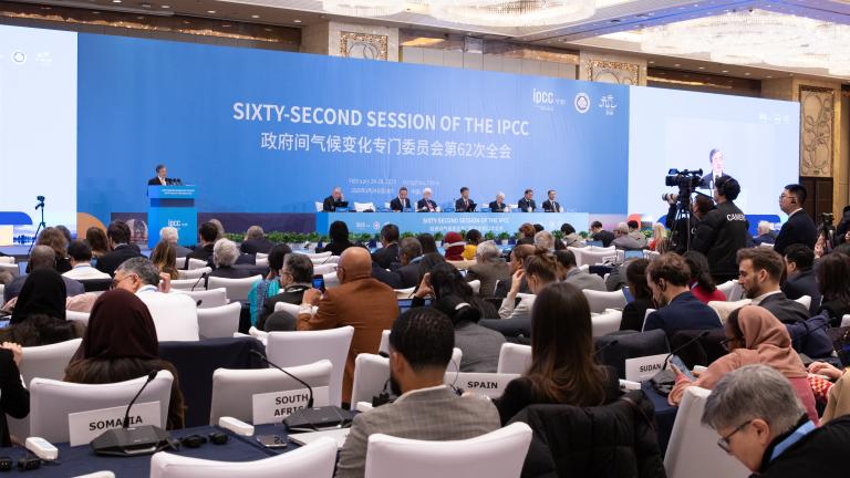 A large conference room with attendees seated. A panel of speakers is at the front beneath a banner reading "Sixty-Second Session of the IPCC." Various country name signs are visible.