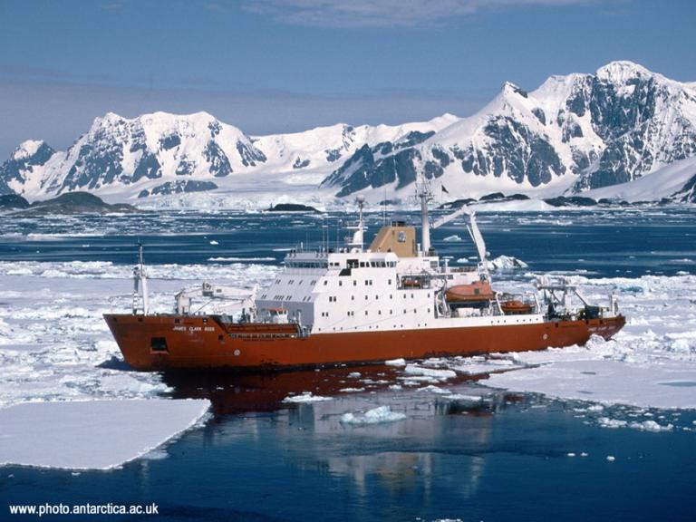 An orange research ship navigates through icy waters with snow-covered mountains in the background.