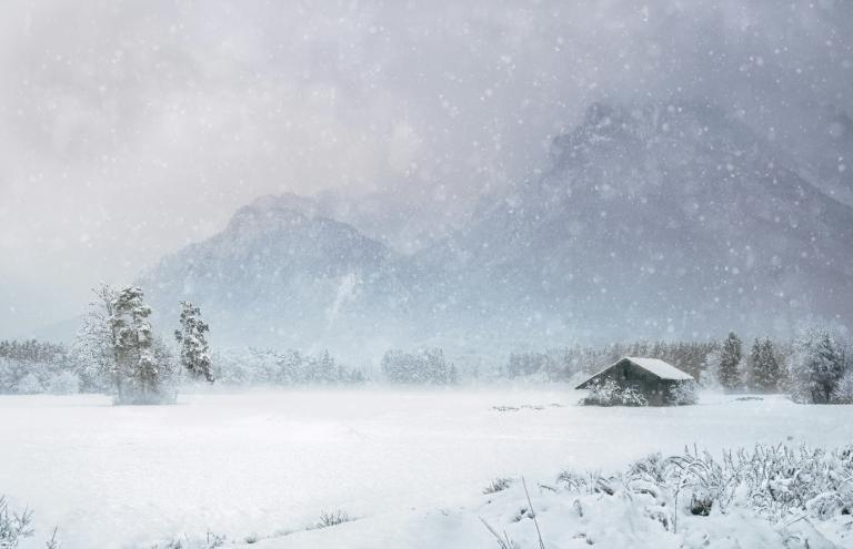 Snow-covered landscape with a small cabin surrounded by trees. Mountains are faintly visible in the foggy, snowy background.