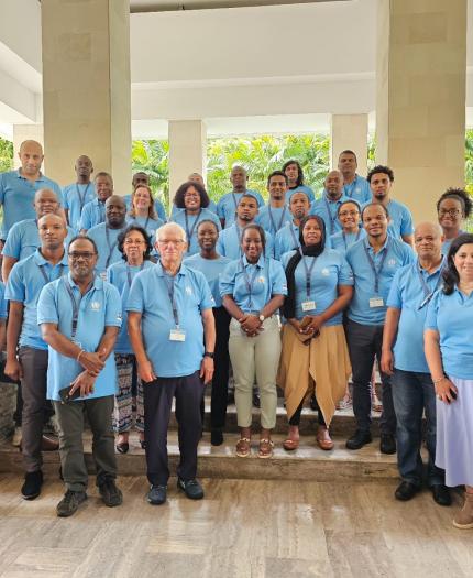 A group of 31 people wearing light blue polo shirts and name badges pose for a photo in a spacious indoor area with columns and greenery visible in the background.