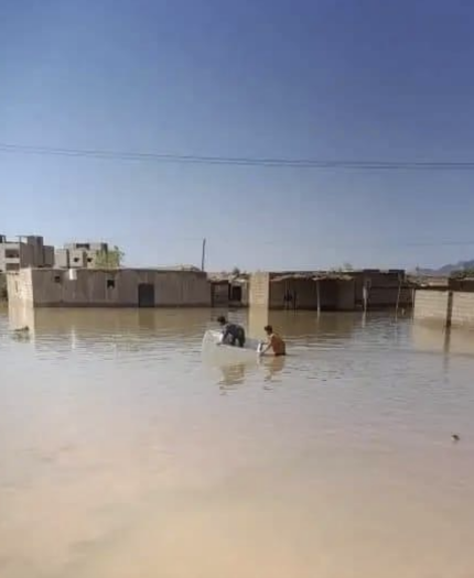 Three people in a small boat navigate through a flooded area with partially submerged buildings under a clear blue sky.