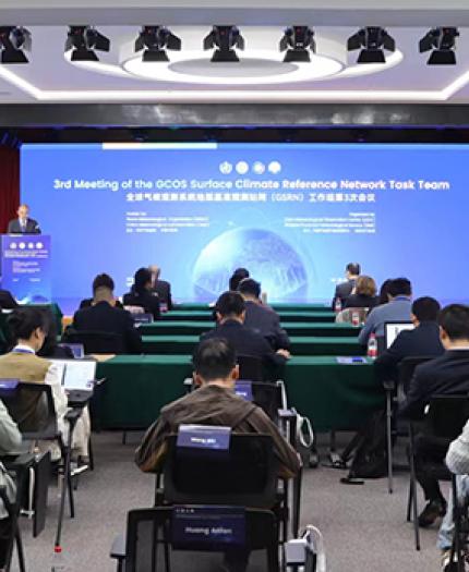 A conference room with attendees viewing a large screen displaying the "3rd Meeting of the GCOS Surface Climate Reference Network Task Team." Chairs and tables are arranged in rows.