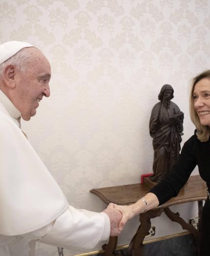 A man in a white robe and skullcap shakes hands with a smiling woman in a black dress in a room with a statue and ornate wallpaper.