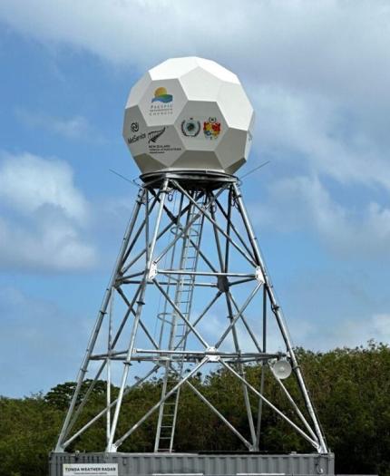 A geodesic dome-shaped weather radar tower stands on a metal framework against a background of blue sky and clouds. Logos are visible on the dome's surface.