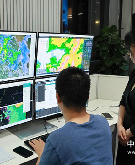 Two people in an office view weather maps and data on multiple monitors in a control room setting.