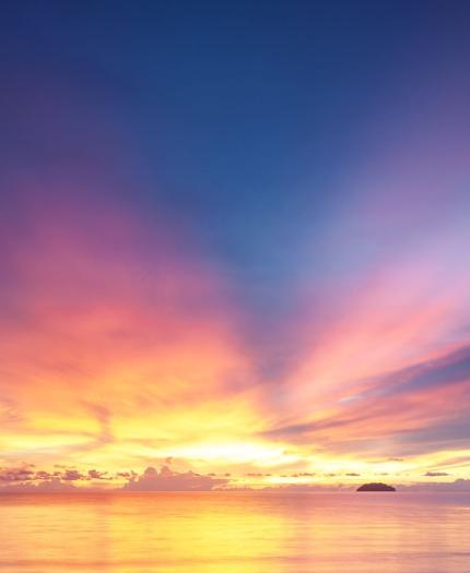 A vibrant sunset over calm waters, with a sky transitioning from orange and yellow near the horizon to deep blue above, and silhouetted islands on the right.