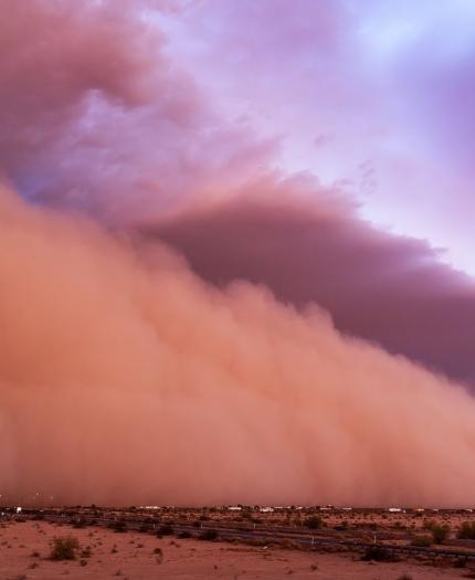 A large dust storm moves across a desert landscape beneath a colorful sky.