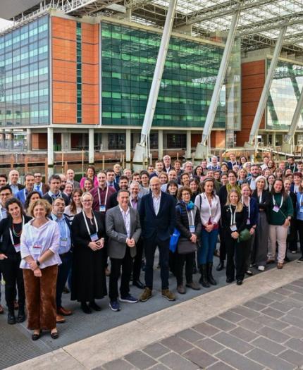 A large group of people pose for a photo outside a modern building with glass and brick elements. They stand on a paved area, some wearing lanyards. The setting appears to be a professional event.