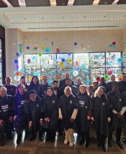 A group of people wearing matching dark outfits pose together indoors in front of a colorful wall display featuring circular designs.