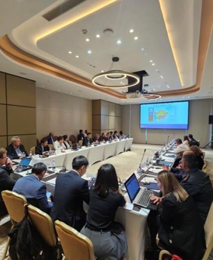 Conference room with a large group of people seated around a U-shaped table. A presentation is displayed on a screen at the front of the room.