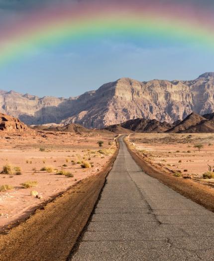 A straight road stretches through a rocky desert landscape with a rainbow arching above distant cliffs under a cloudy sky.