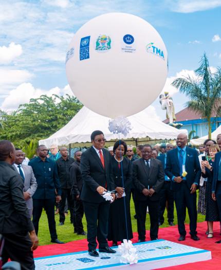 A group of people, including officials in suits, stand on a red carpet outdoors beneath a large white balloon with logos, surrounded by greenery and tents.