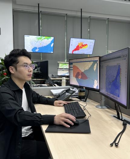 A person sits at a desk in an office with multiple monitors displaying weather maps and data.