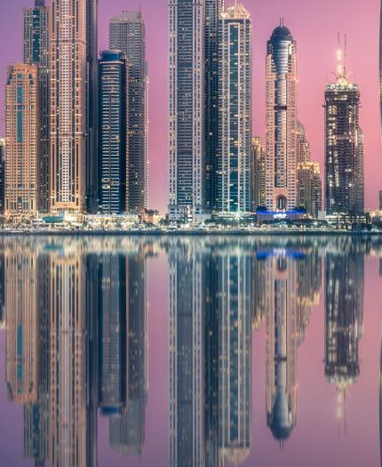 City skyline with tall, illuminated skyscrapers reflecting in a calm body of water during twilight. The sky is a gradient of pink and purple hues.