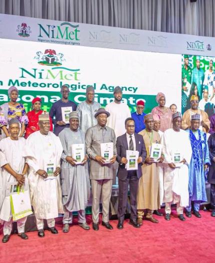 A group of people, mostly men, stand on a stage holding booklets. The banner behind reads "Nigerian Meteorological Agency" with "Seasonal Climate" partially visible.