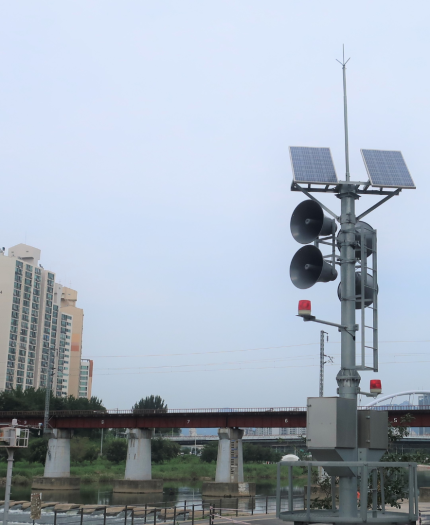 A tall pole with solar panels and loudspeakers stands by a river, near a railway bridge and high-rise buildings under a cloudy sky.